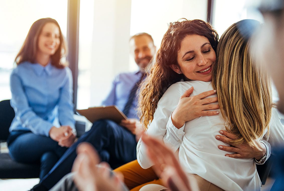 Two woman embraces lovingly as others watch and smile during group therapy