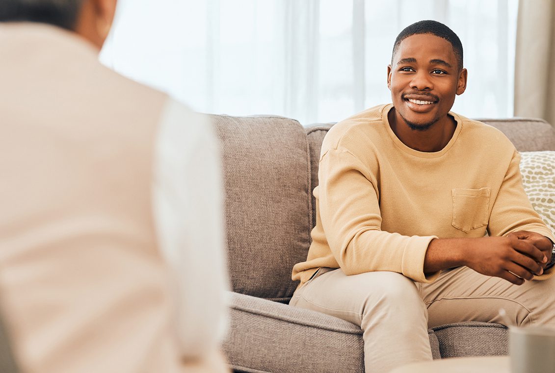 African American man smiles while leaning forward on the sofa to listen to his partially unseen therapist