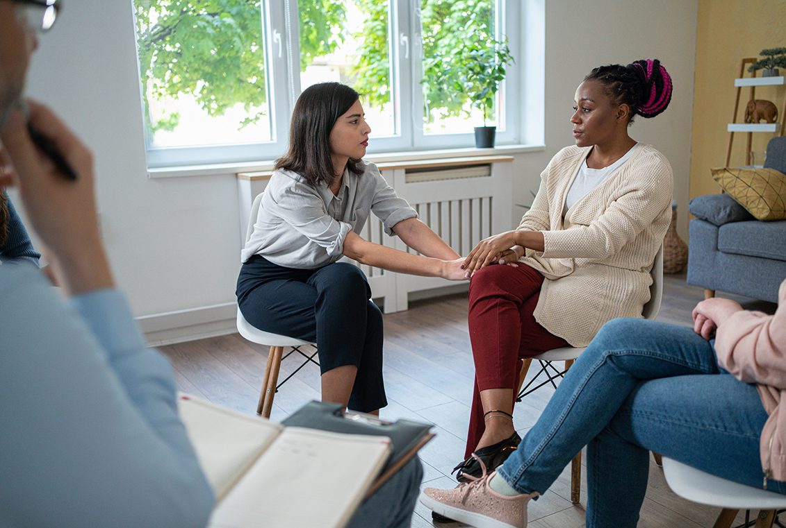 Woman reaches to hold friend's hand during a group therapy session as others watch