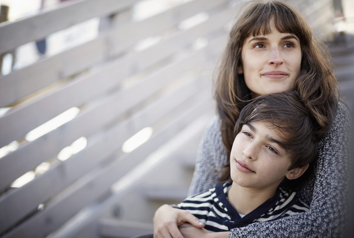 Mother hugs her teenage son from behind as they sit on steps together
